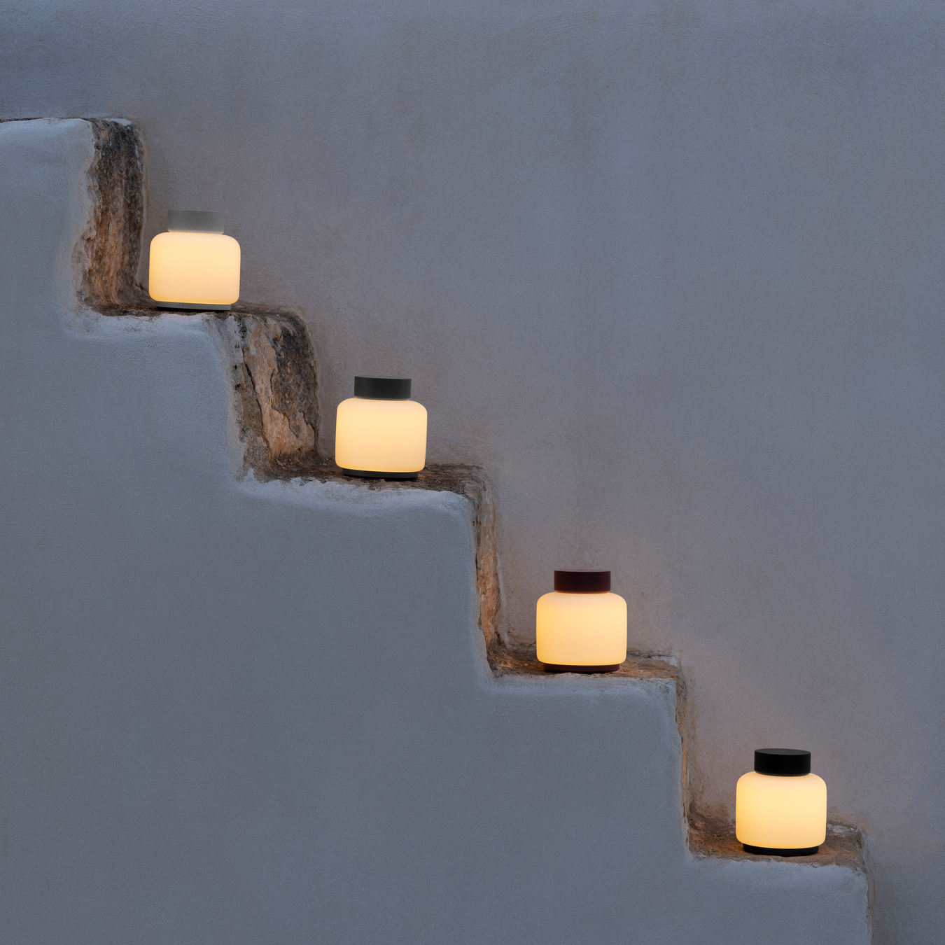 Four FAWN Lanterns sitting on stairs of a minimalist building all lit up with warm color lighting.