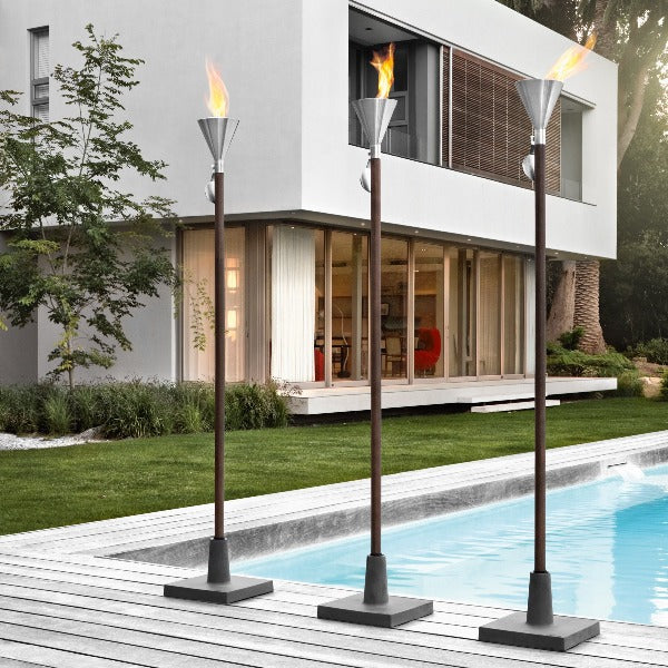Three outdoor torches with flames lit in front of a modern house with a pool.