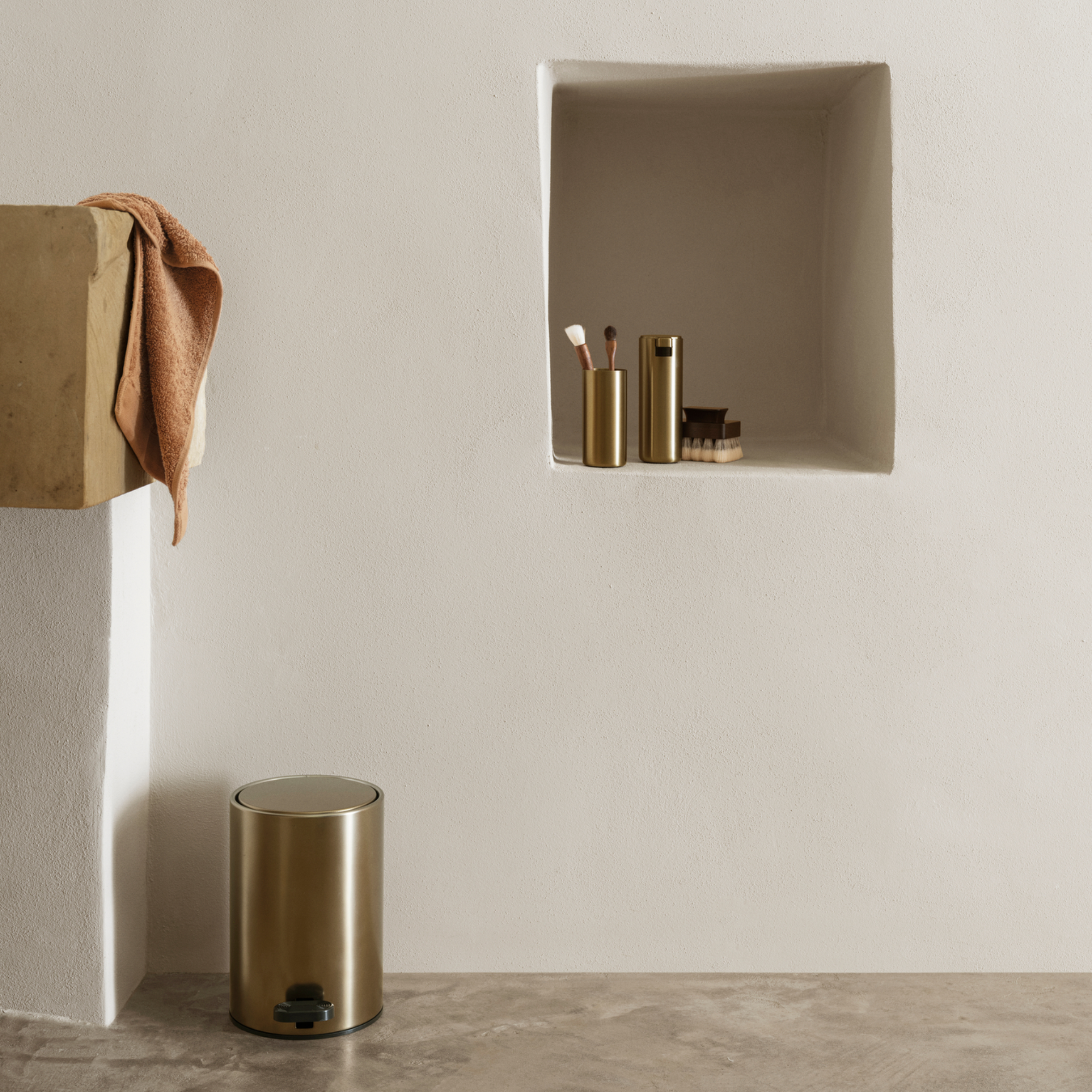 MODO Brass Wastebasket next to a shelf with MODO Tumbler and Soap Dispenser along with grooming brush on a neutral background.