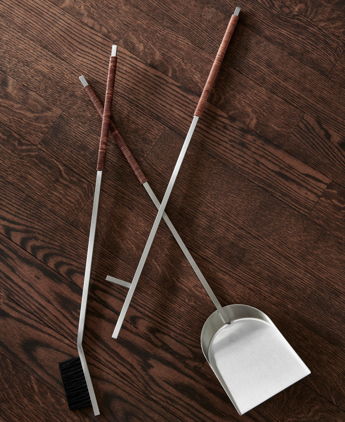 Fireplace tool set with long silver handles, leather wrapped grips, a black brush bristle head, and a stainless dustpan, arranged on a dark wood floor.