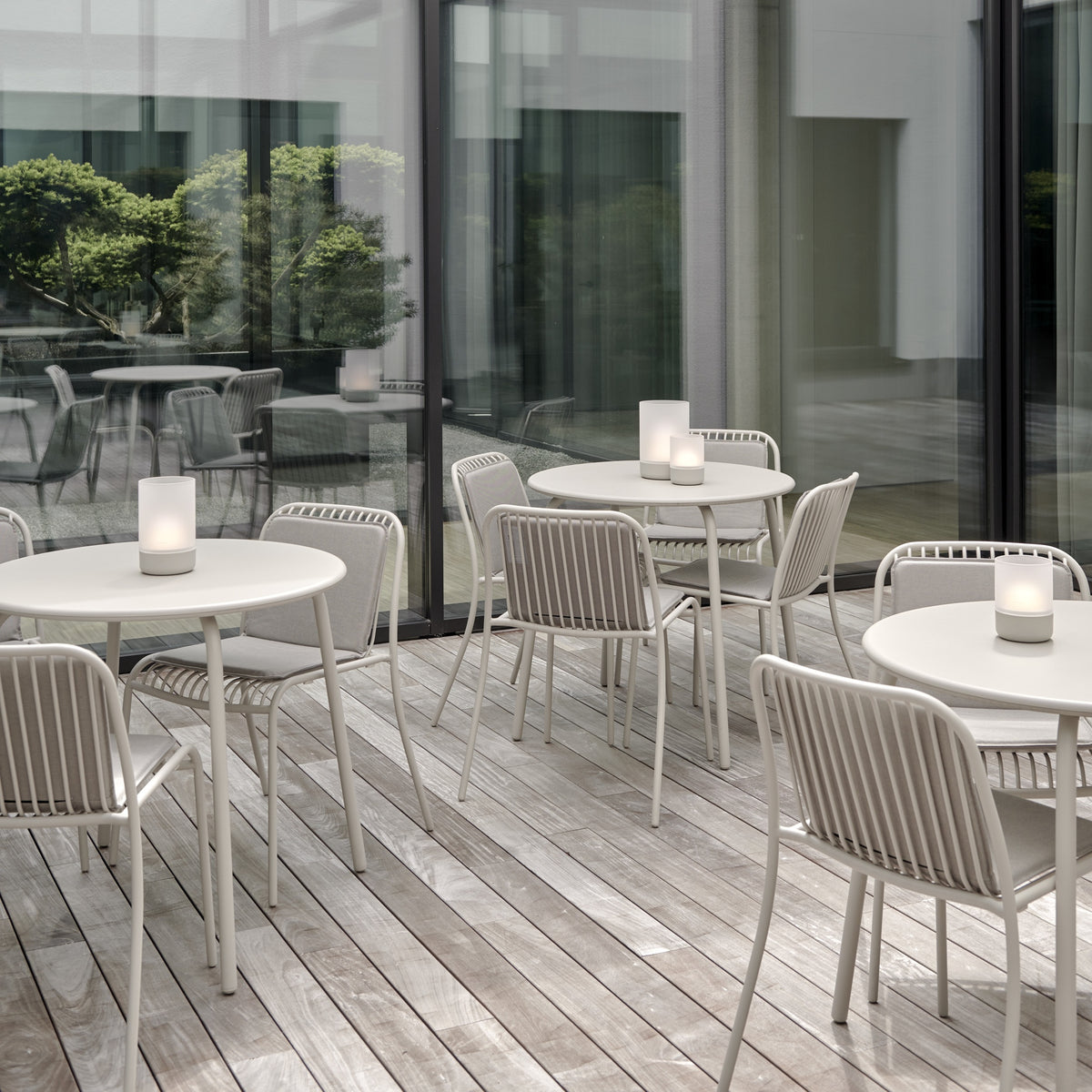 Outdoor patio dining set with round white tables and light gray woven sling chairs, arranged on a weathered wood deck in front of large glass windows.