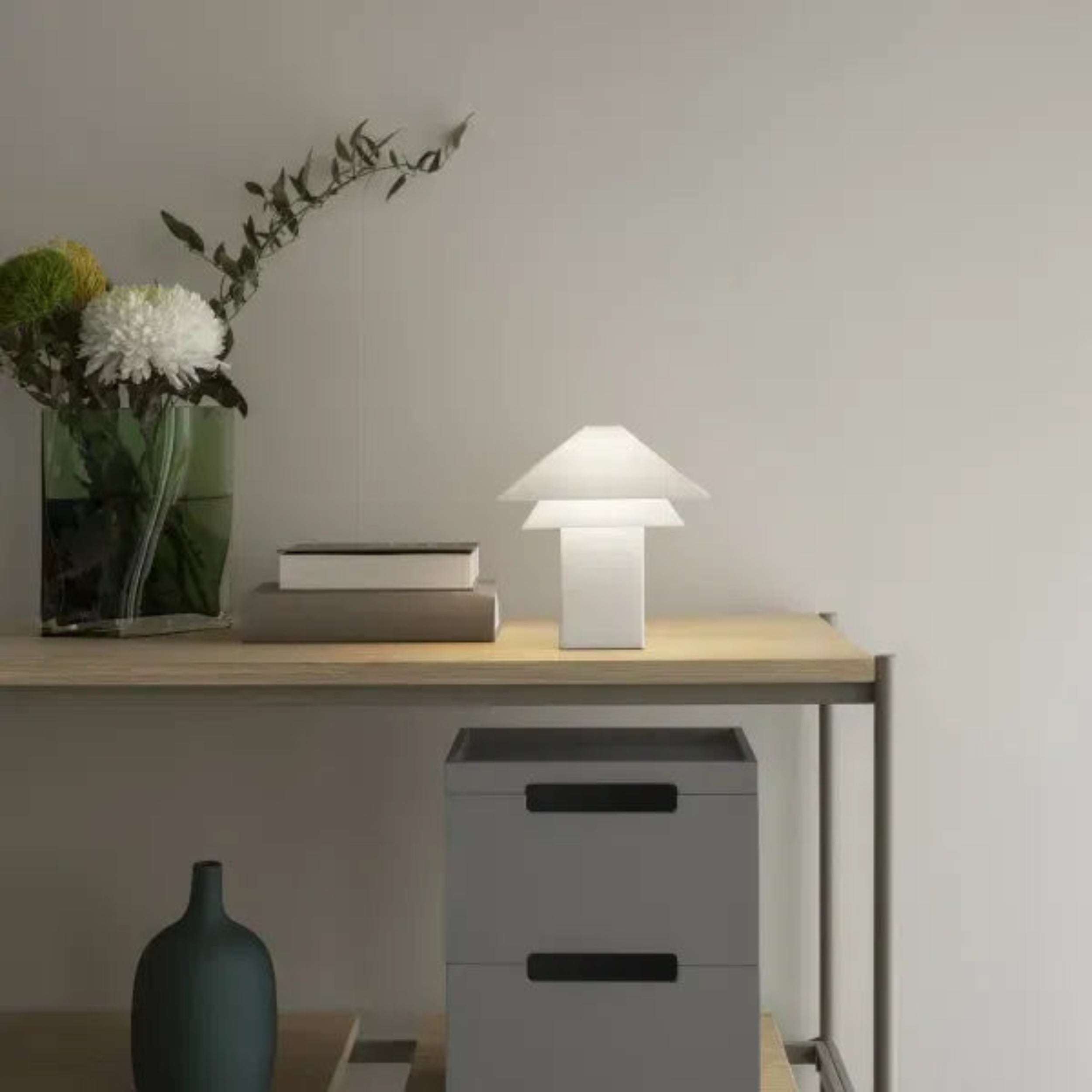 Modern desk setup with a lamp, vase, and books against a neutral wall.
