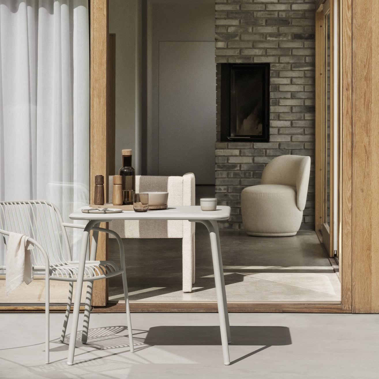 Outdoor patio setting with bright sunlight focusing on a square metal table set for a meal with wire back chair and a view of modern living room through the open sliding door