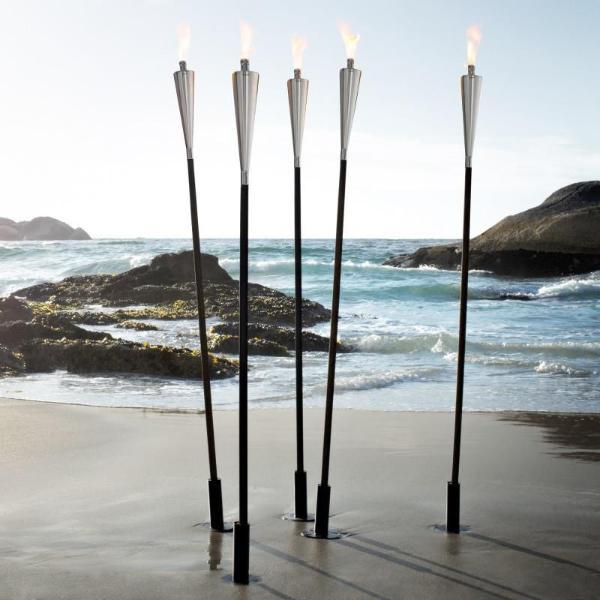 Outdoor garden torches with stakes planted in the sand next to roaring waves and rocks on the beach