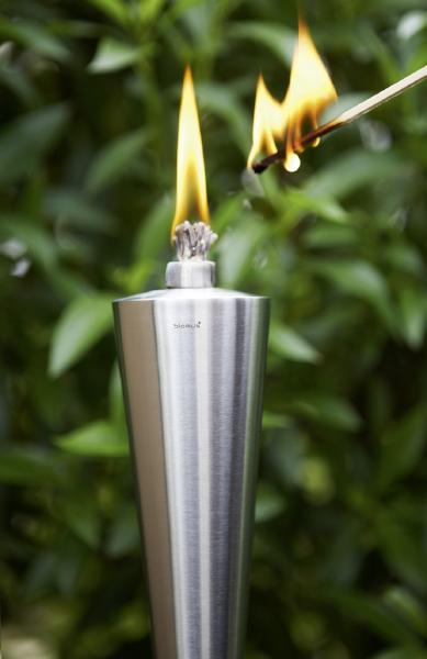 Stainless steel garden torch being lit by a large match in a lush green outdoor setting