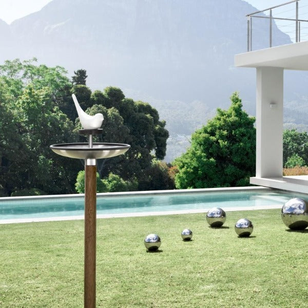 Stainless steel bird feeder on a wooden stake next to a pool and silver balls in a modern backyard
