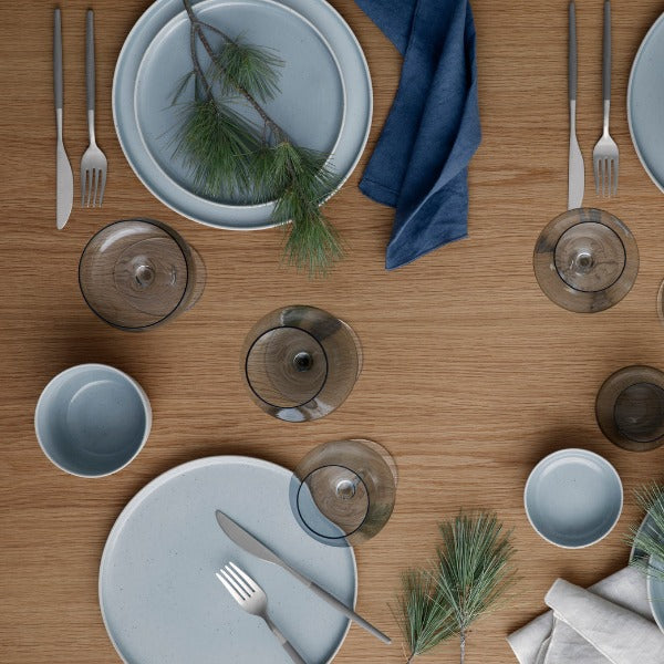 Blomus SABLO dinnerware in Stone gray arranged on a wooden dining table with glassware, silver cutlery, blue napkins, and pine greenery accents