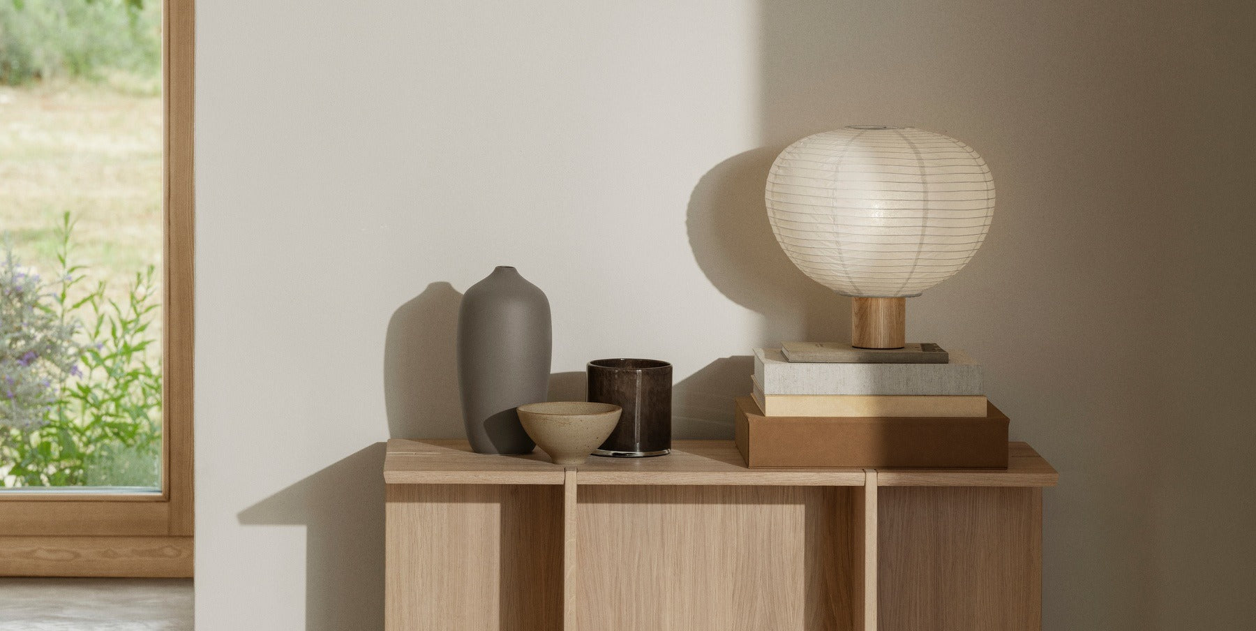 Minimalist wooden blomus sideboard by a window, styled with neutral-tone ceramic vases, stacked books, and a blomus paper lantern lamp, bathed in soft natural light.