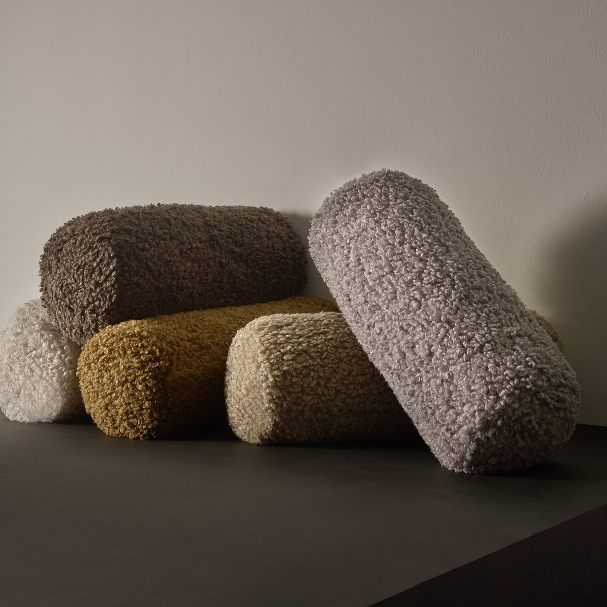 TEDDY Round Bolster Accent Pillows stacked on a shelf in 5 colors with a grey background