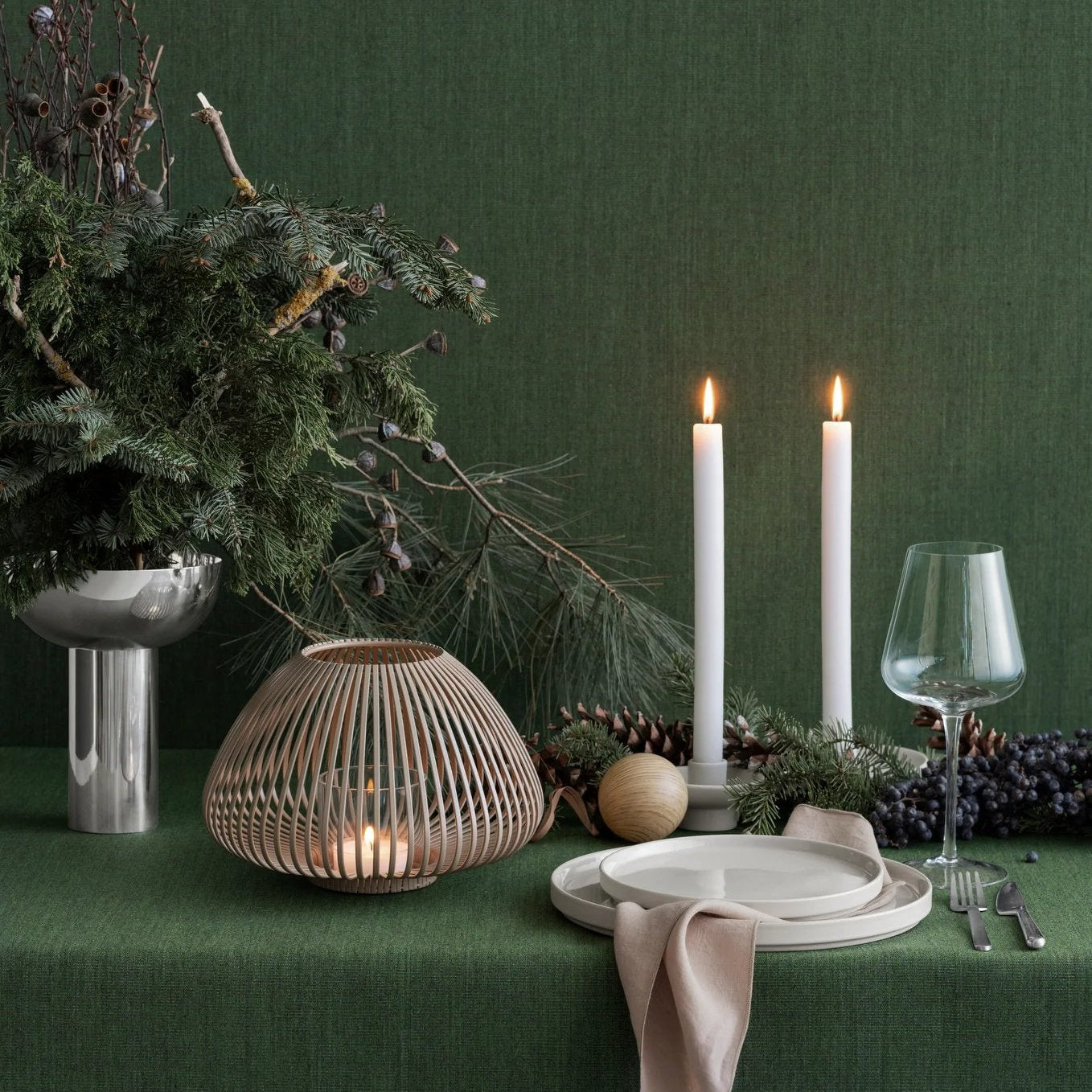 Decorative table setting with candles, greenery, and a woven lantern against a dark green wall.