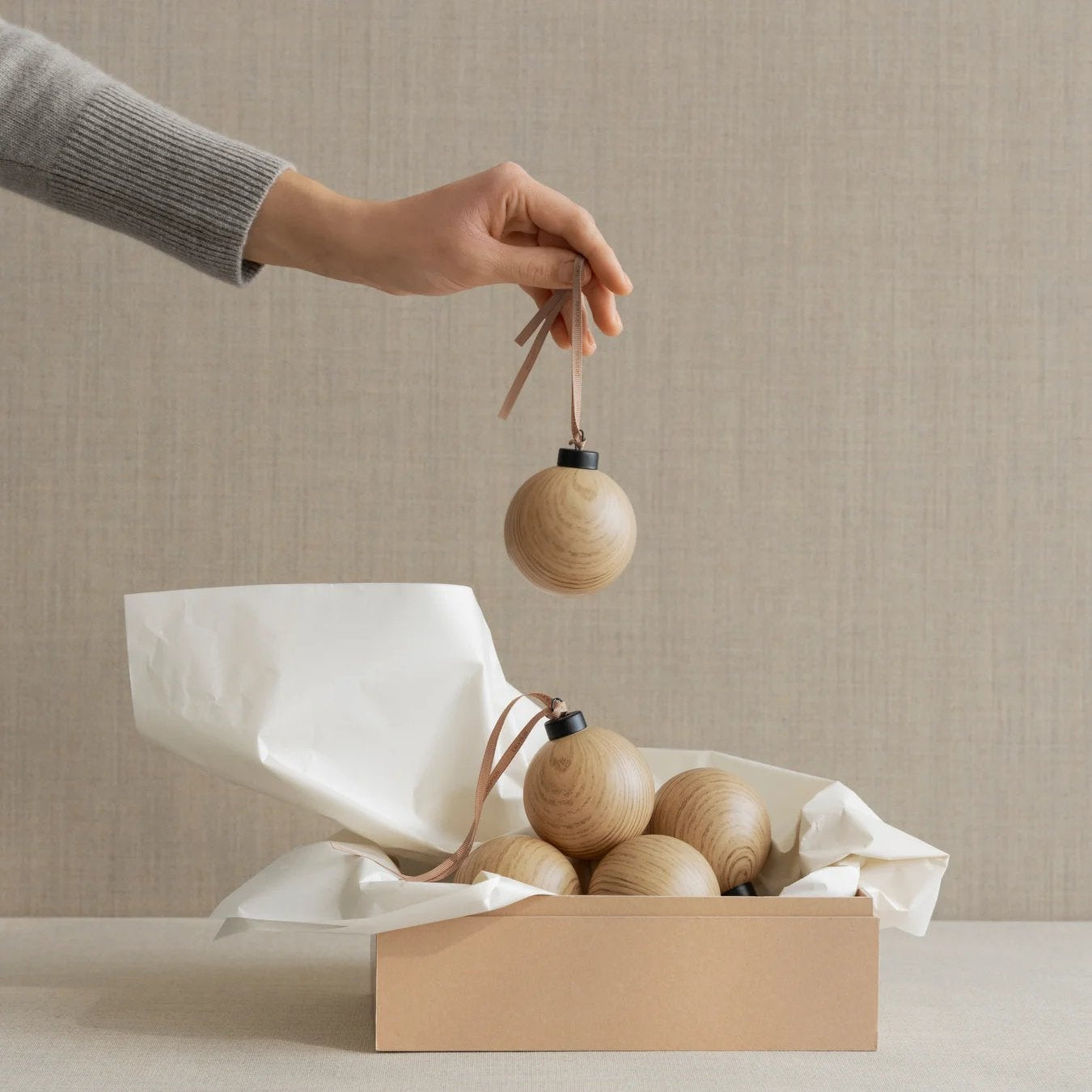 Person holding a wooden ornament above a box with more ornaments on a neutral background