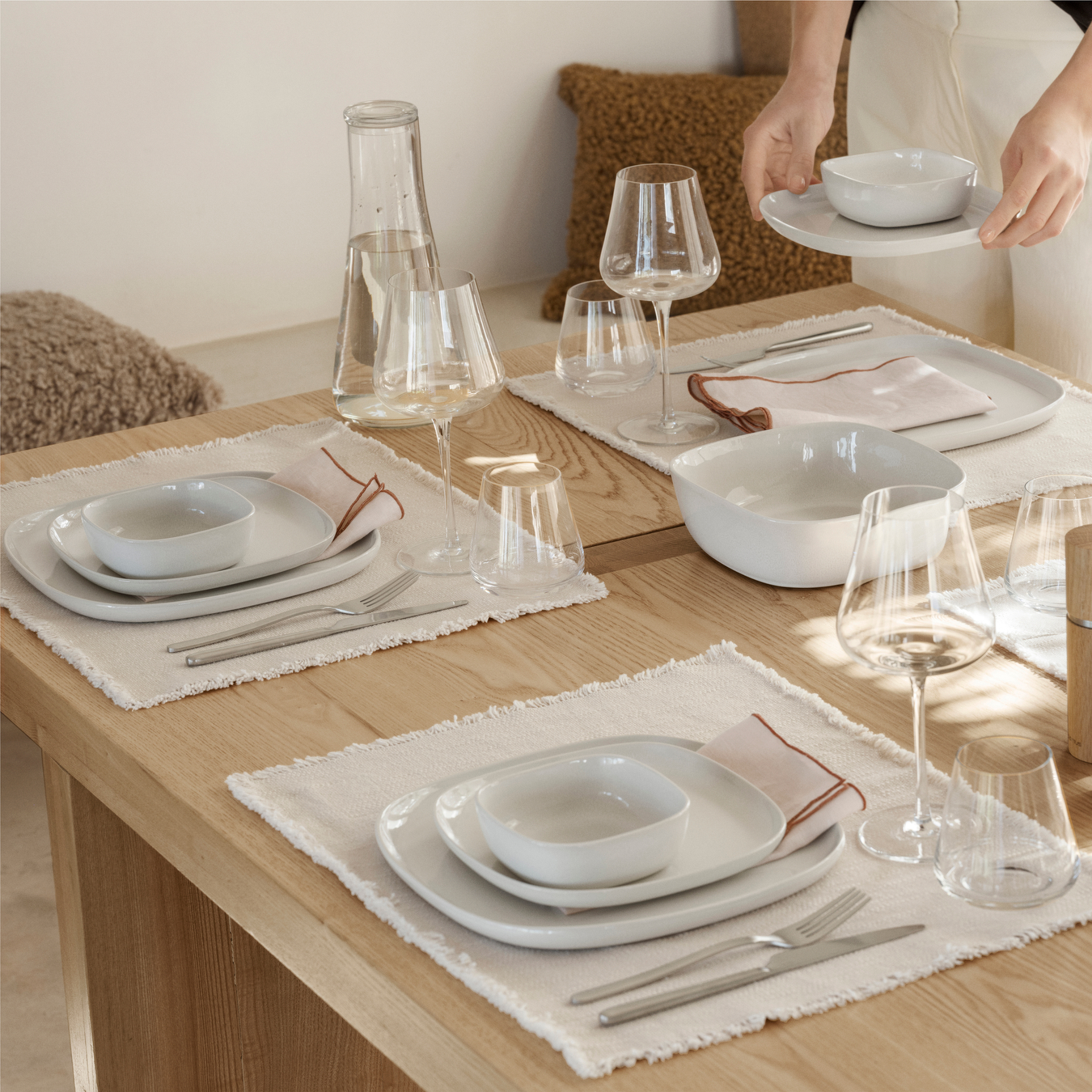 VAHLO Serving Bowl and place settings in Moonbeam color on a table with BELO Stemware and and silverware. Female hands shown finishing table setting.