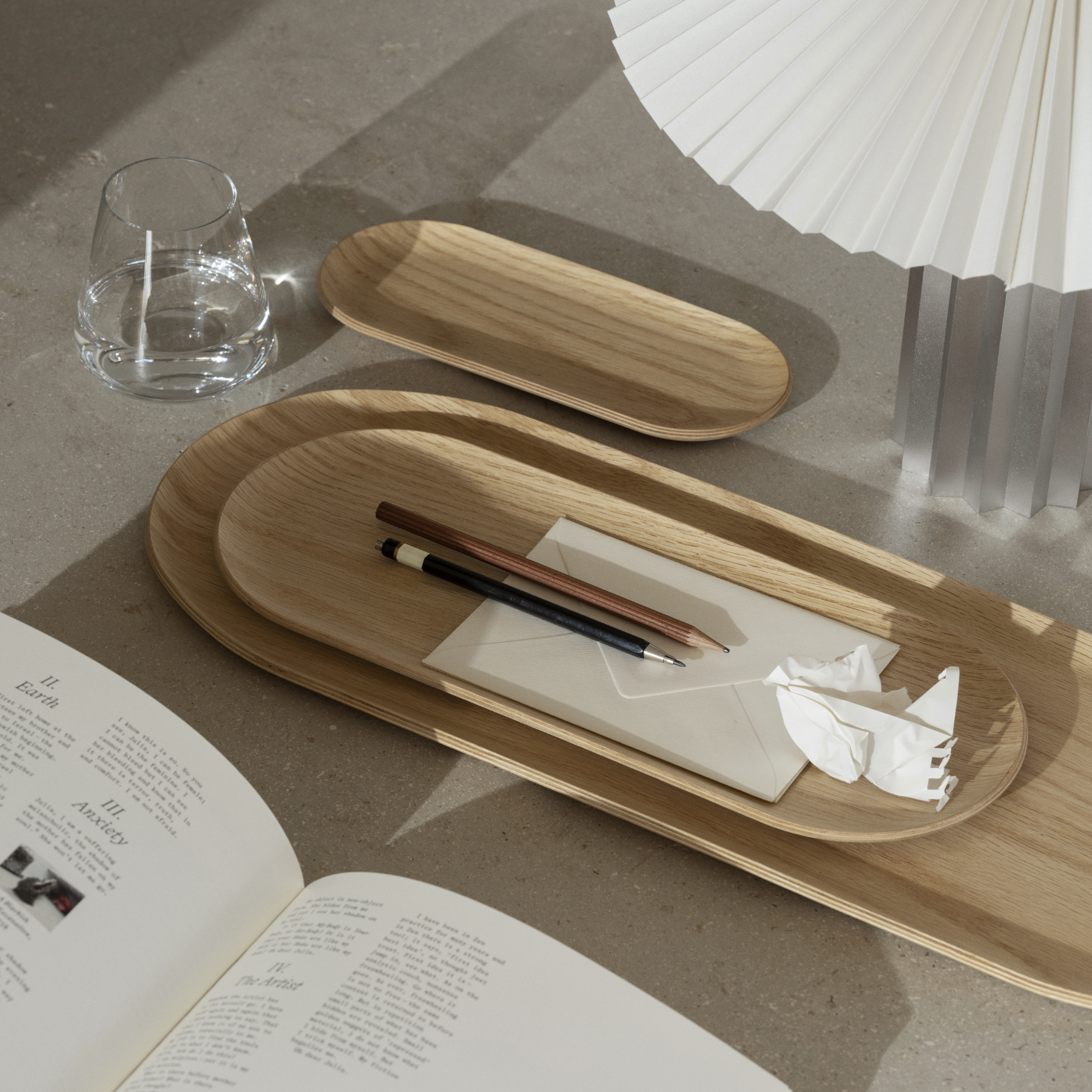 Three sizes of oak BAKI Trays placed in an office type setting showing an open book, pencil and pen, letter and lamp.