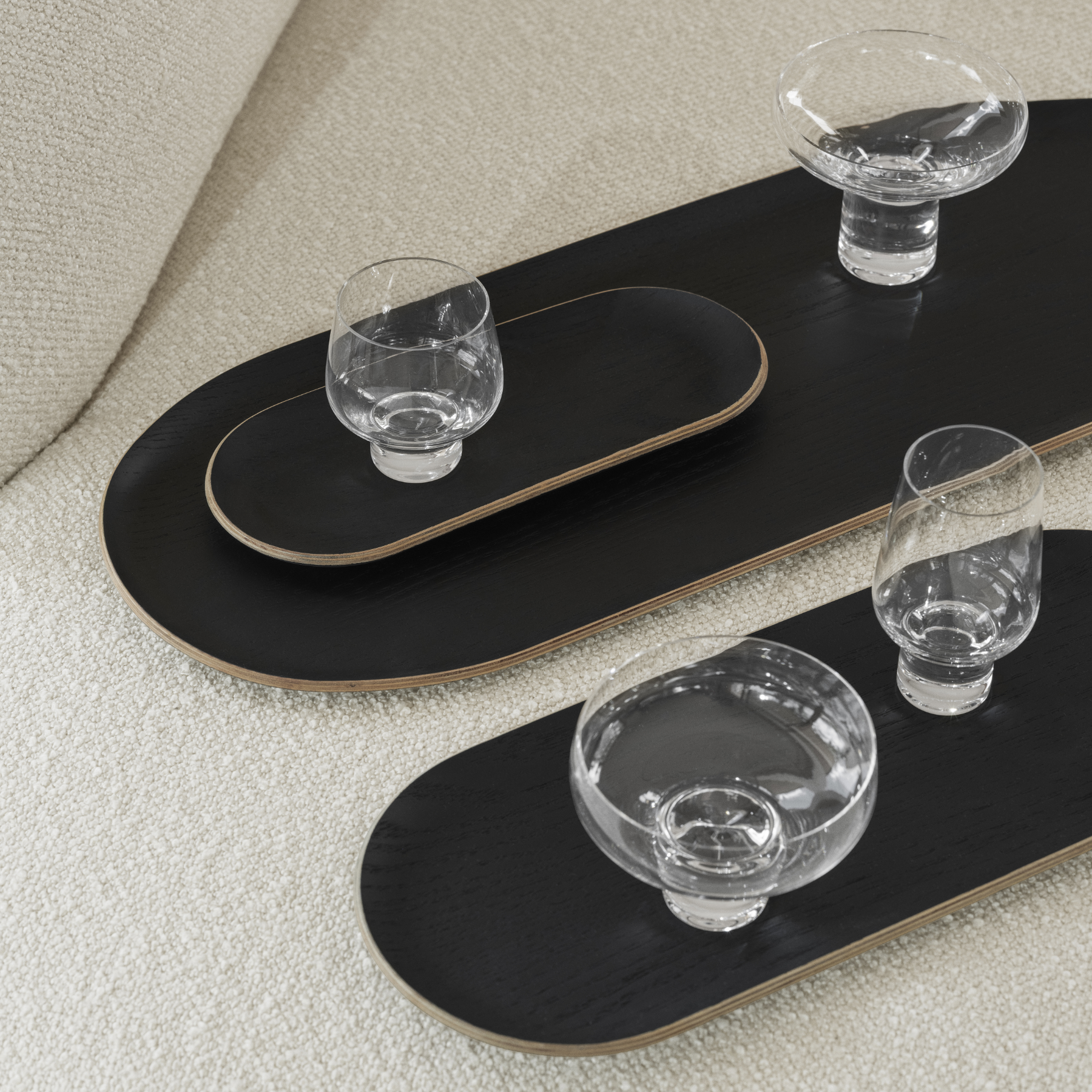 BAKI Black Trays in three sizes holding KOYOI Glassware placed on a neutral colored sofa. 