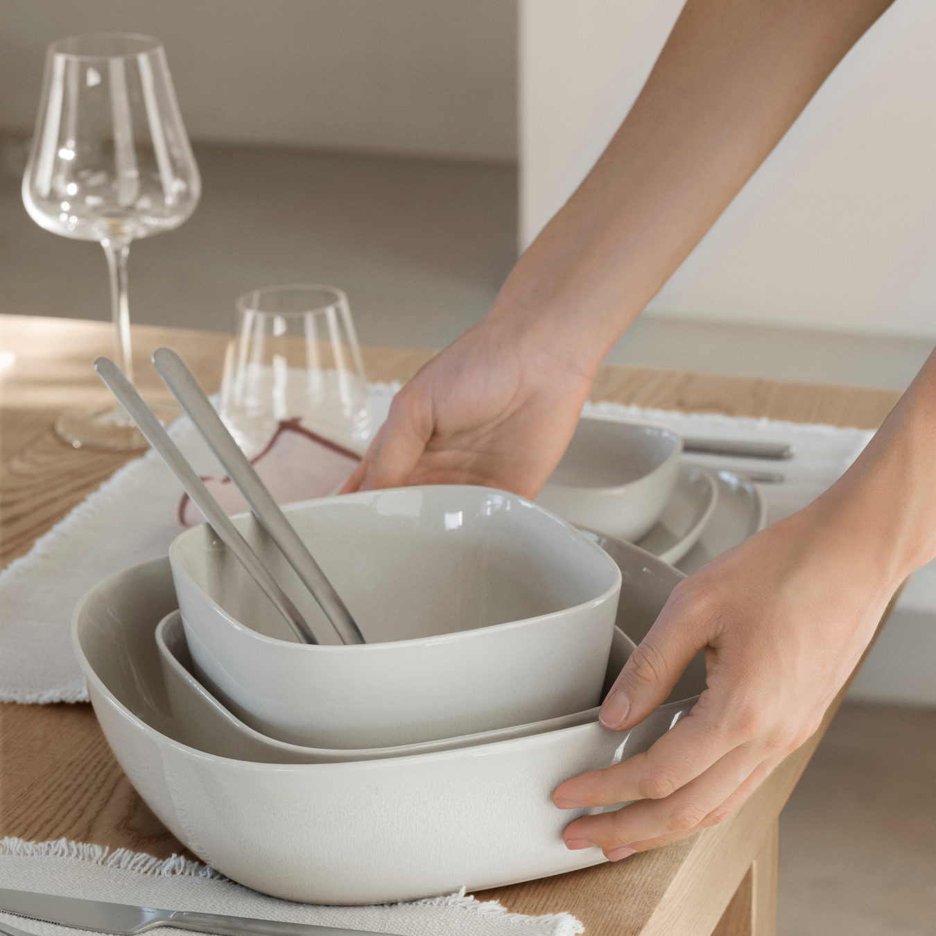 VAHLO Serving Bowl and Salad Bowls being placed on a set wood table by a female. Table is set with VAHLO Moonbeam dishes and BELO Stemware.