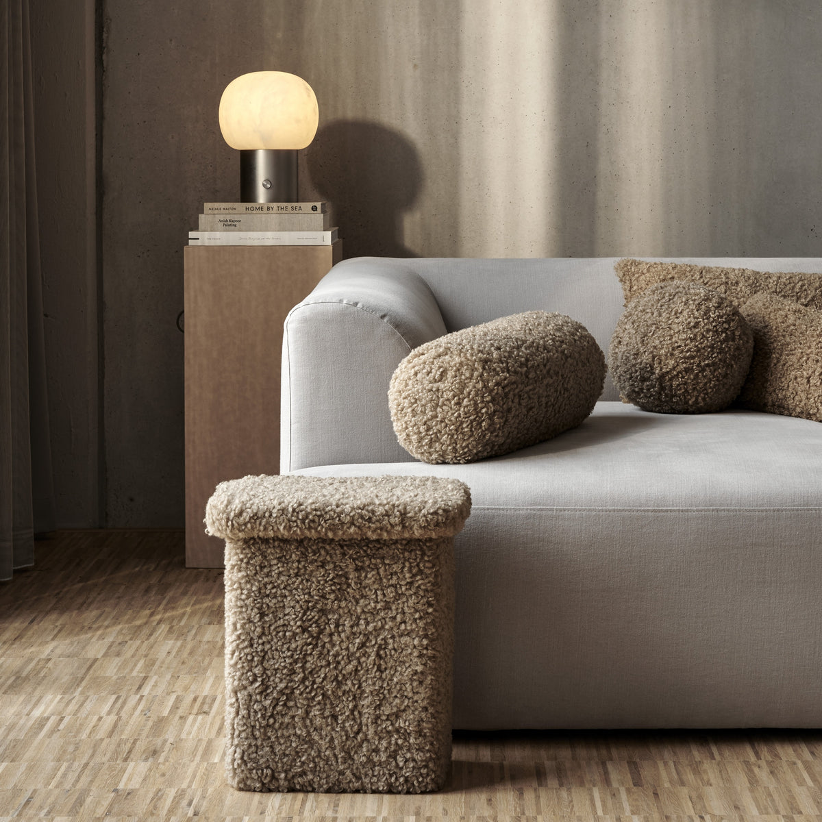 A modern light gray sofa in a styled living room setting, featuring textured TEDDY Pillows and a matching textured ottoman, with a side table and glowing lamp in the background.