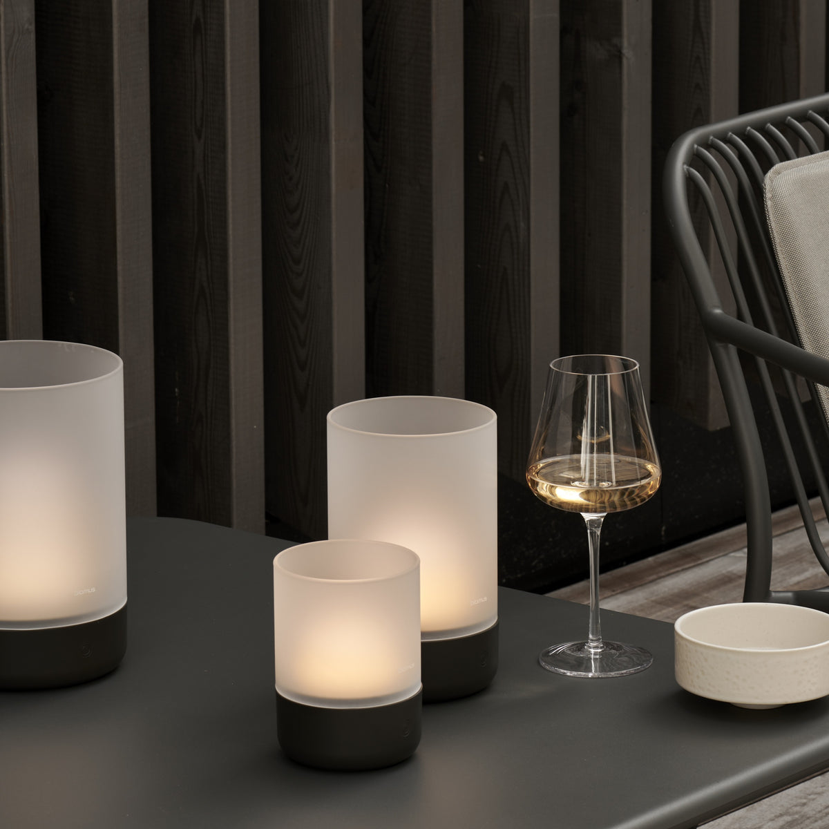Three modern cylindrical LED CALMA lamps with frosted white hurricanes and granite grey bases are arranged on a dark table, alongside a wine glass, a small bowl, and a chair in a patio setting.