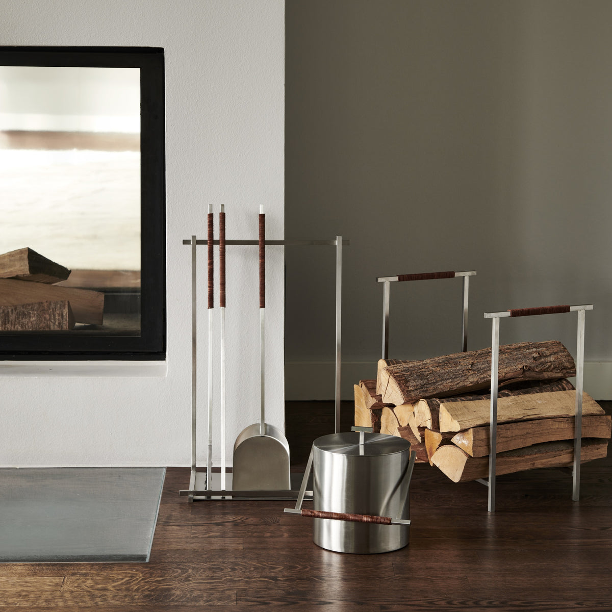 A stainless steel fireplace tool set with a bucket, shovel, and poker stands on a dark wood floor next to a stacked wood firewood rack in a modern room setting.