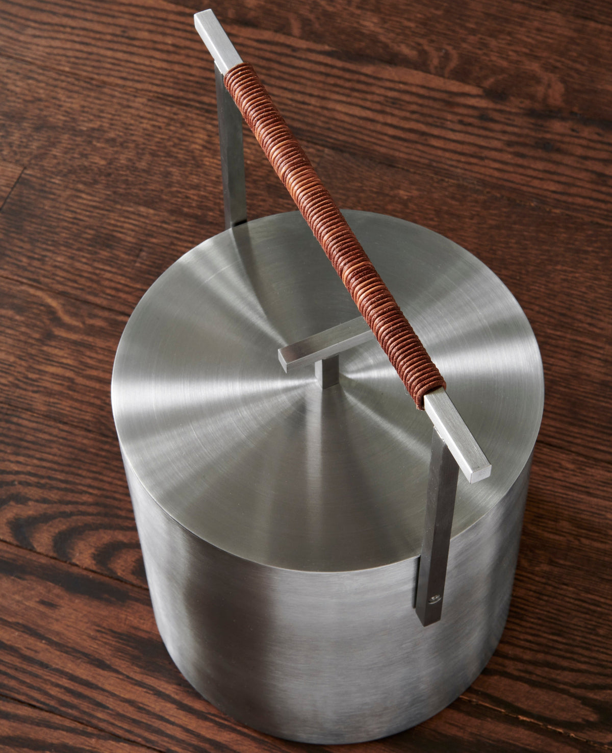 A brushed stainless steel ash bucket with a lid, featuring a handle wrapped in leather cord photographed on a dark wood surface.