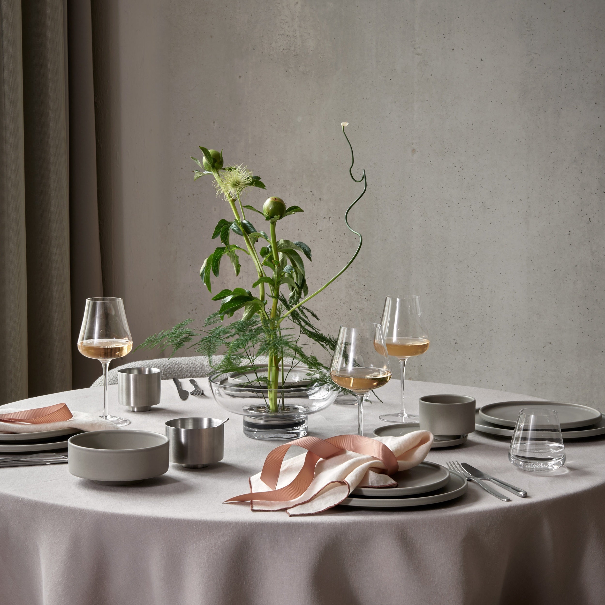 A styled dining table set with ceramic plates, bowls, and small metal cups, paired with blush pink napkins and wine glasses, centered around a green floral centerpiece against a textured gray wall.