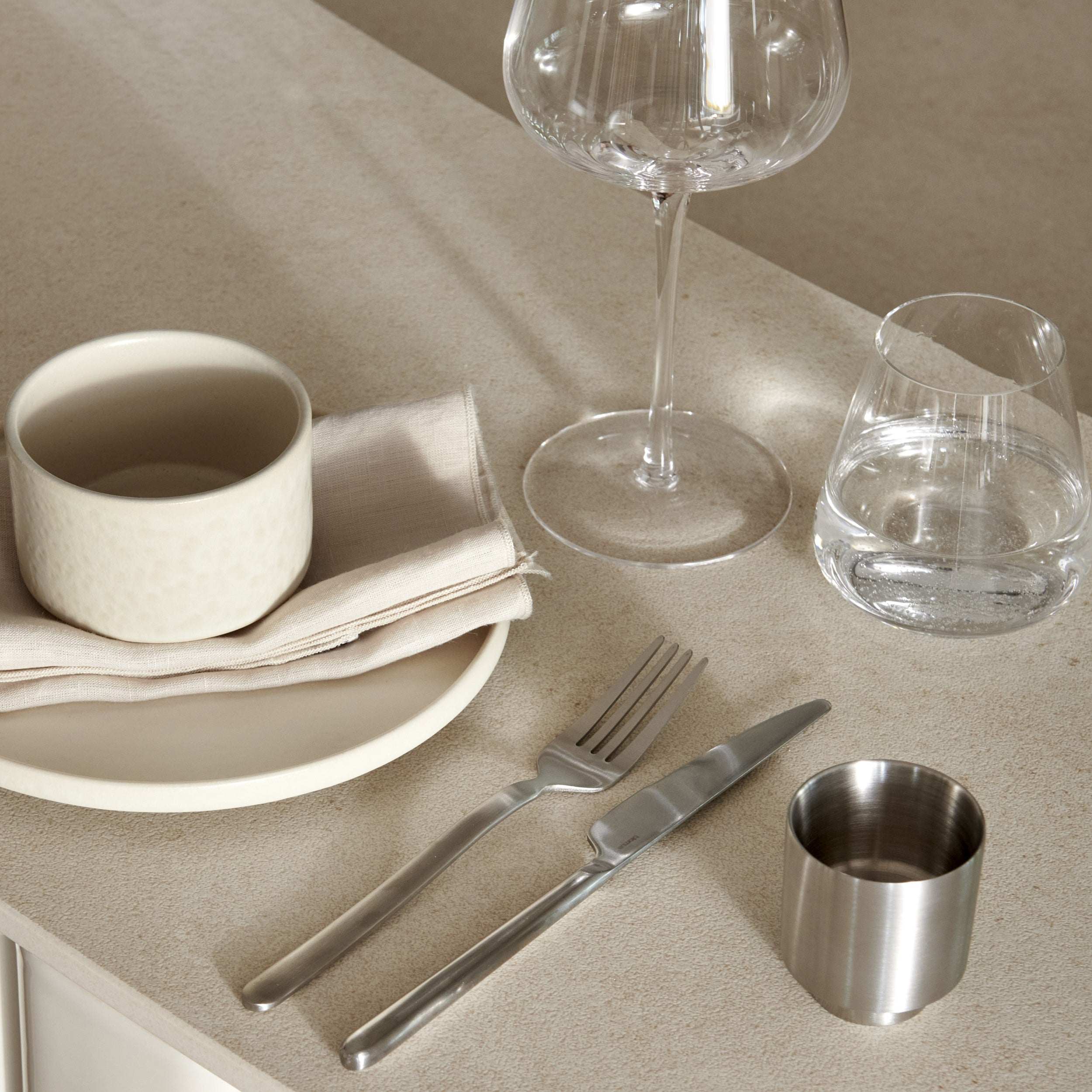 A styled tabletop dining scene with a cream ceramic cup and plate, folded napkin, silver fork and knife, a small stainless steel cup, a wine glass, and a tumbler on a beige surface.