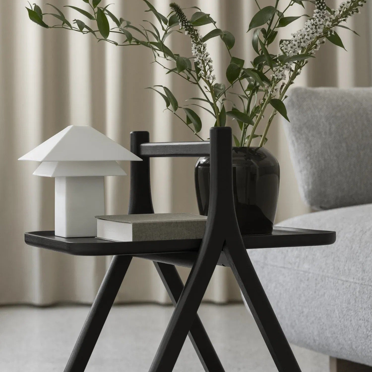 Small black side table with a white lamp, book, and vase with greenery in a room setting.