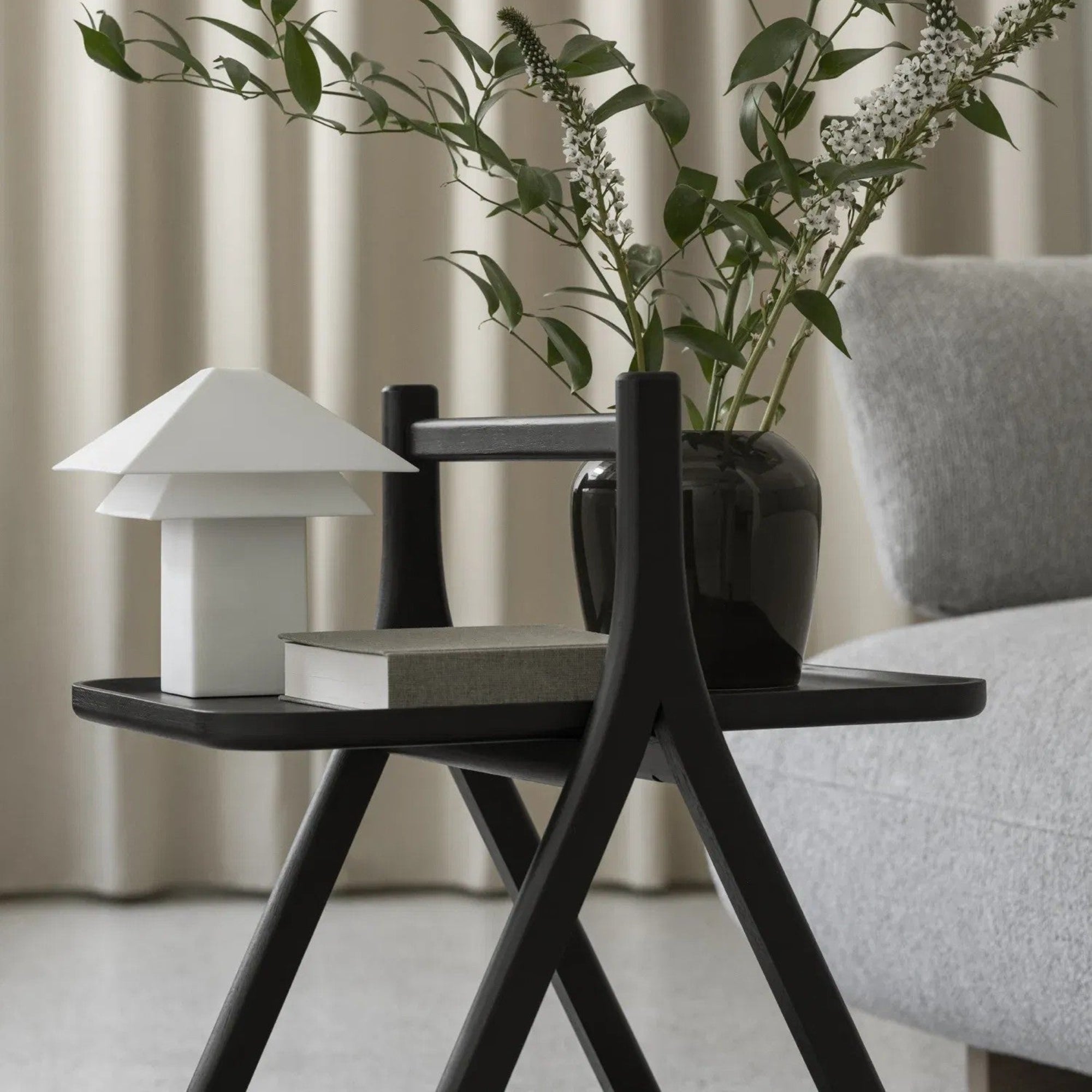 Small black side table with a white lamp, book, and vase with greenery in a room setting.
