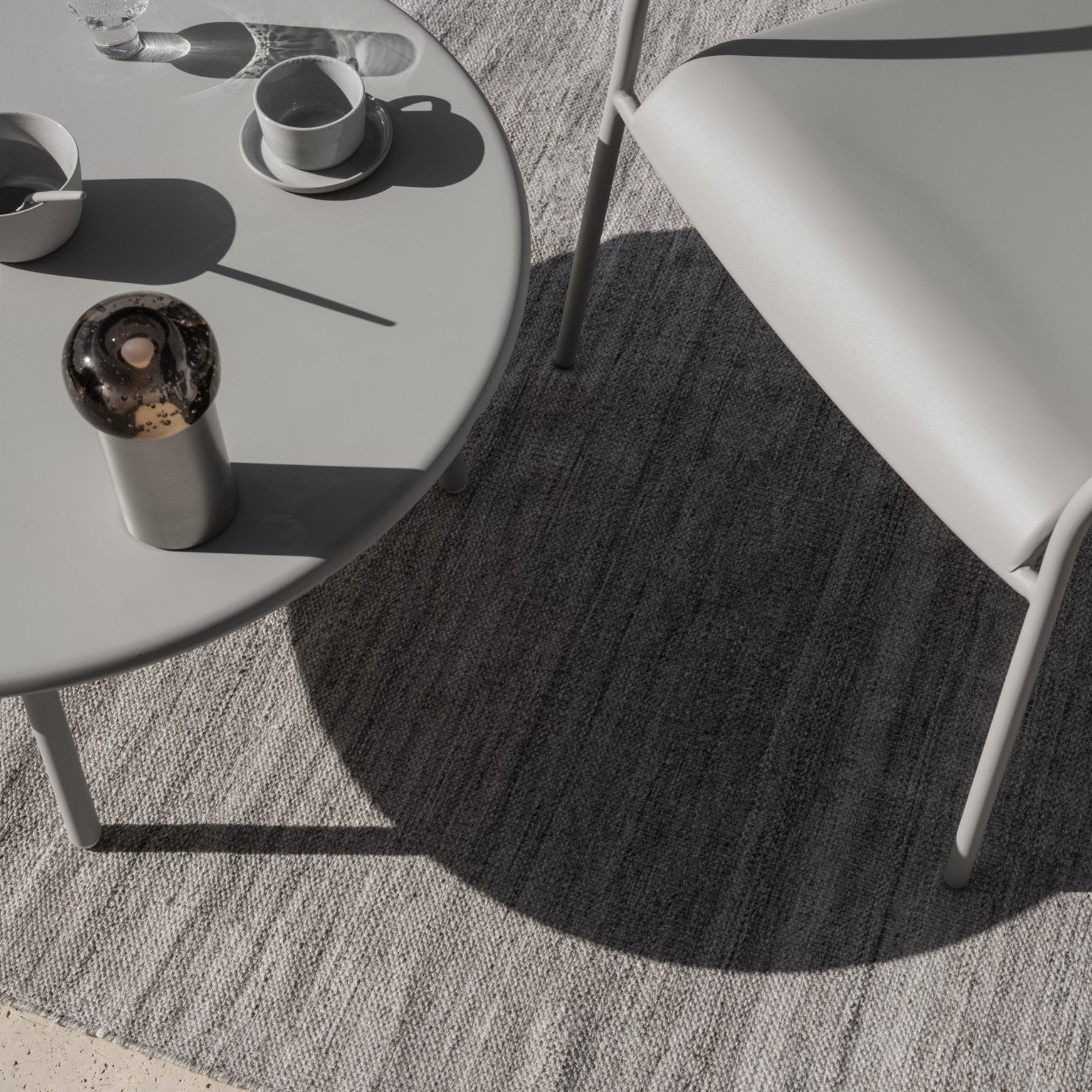Looking down on an outdoor table and chair placed on a rug. A lamp and coffee cup are on the table with heavy shadows from the morning sun
