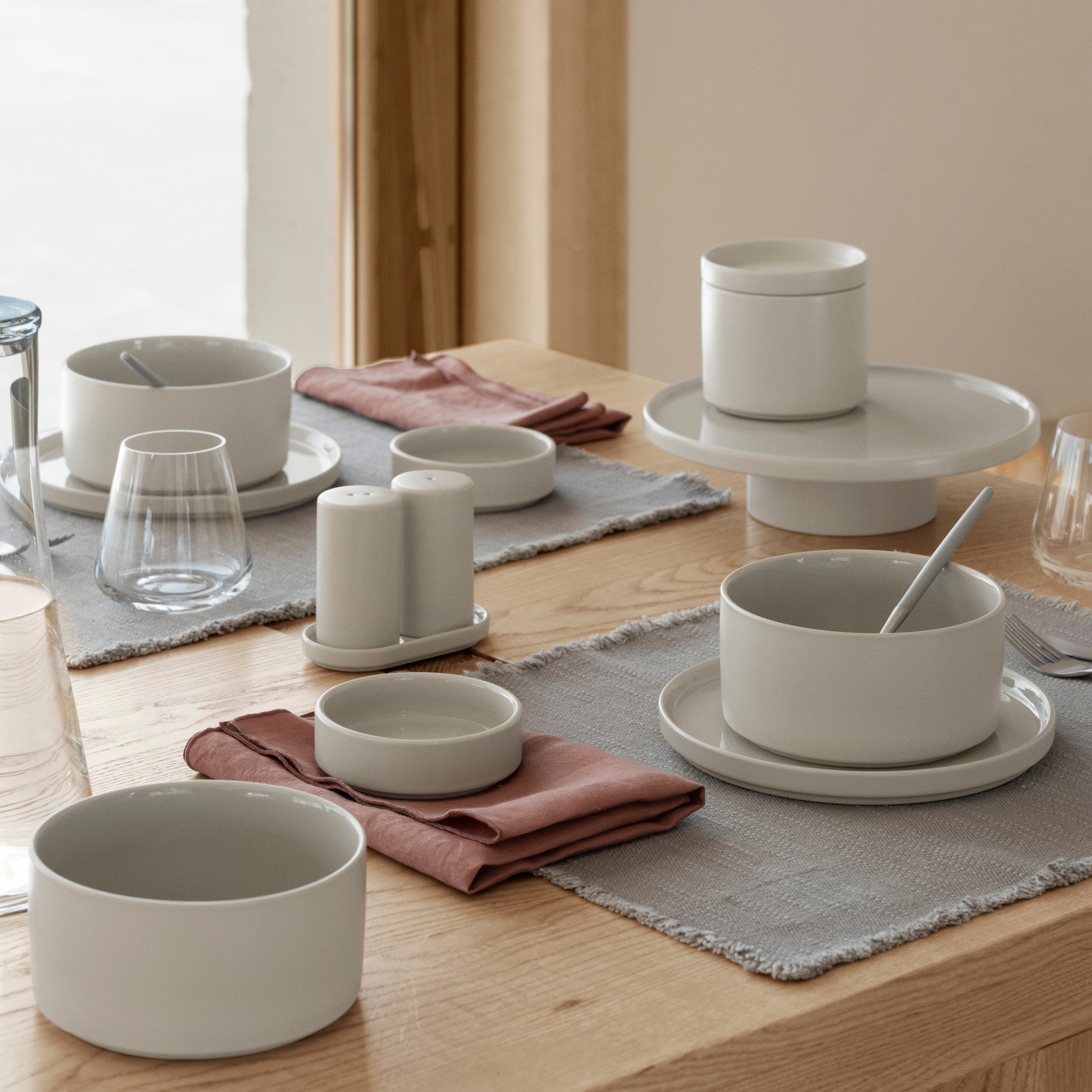 PILAR Dishes set on a wood table including plates, bowls, canisters and salt and pepper set with placemats, silverware and BELO Stemware. et of white ceramic bowls and cups on a wooden table with gray placemats.