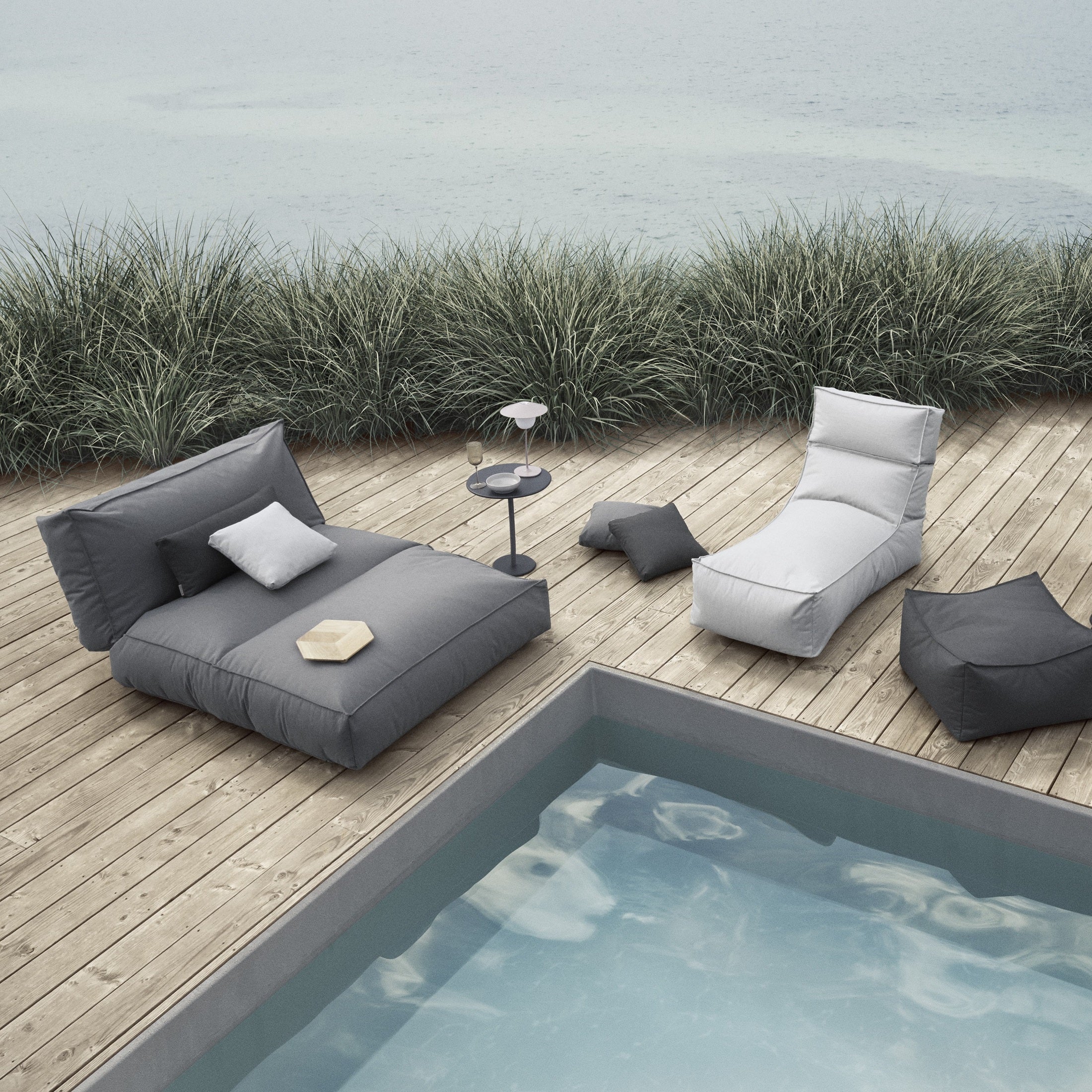 oversized outdoor lounger with side table on a wooden pool deck next to tall grass