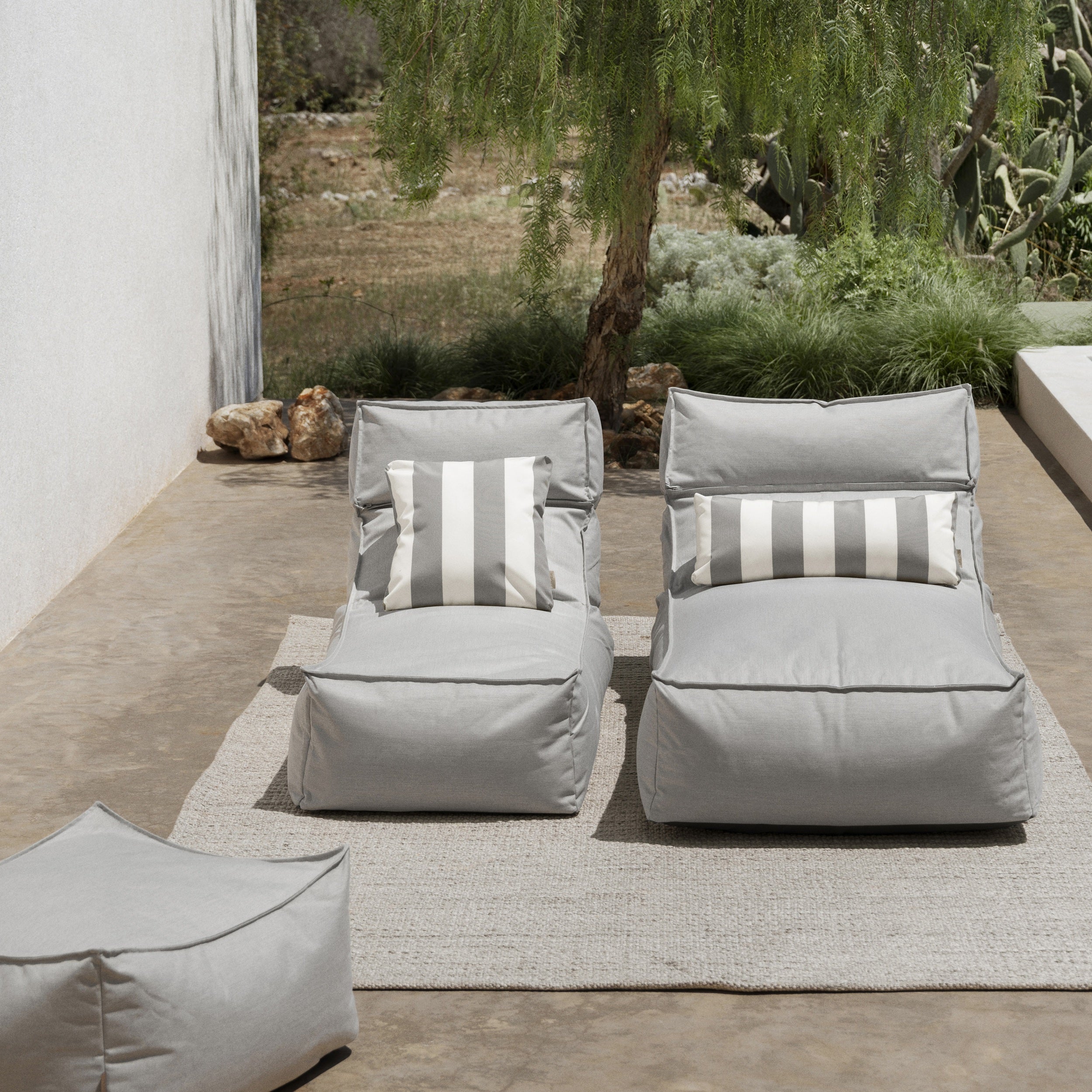 Two gray outdoor lounge chairs with striped cushions on a patio with plants and rocks in the background.